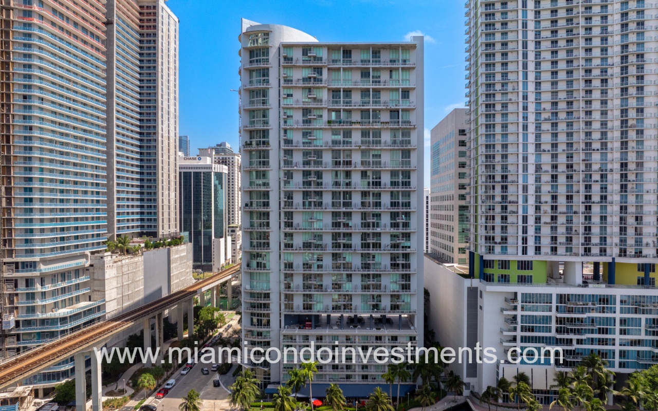 Skyline of tall modern Miami condos along a street with palm trees and an elevated rail line nearby.