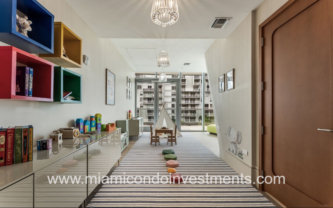 UNA Residences condominium building bright children’s playroom with colorful wall shelves, toys, books, a small table and chairs, and a striped rug in a sunny space for kids age 2–6.