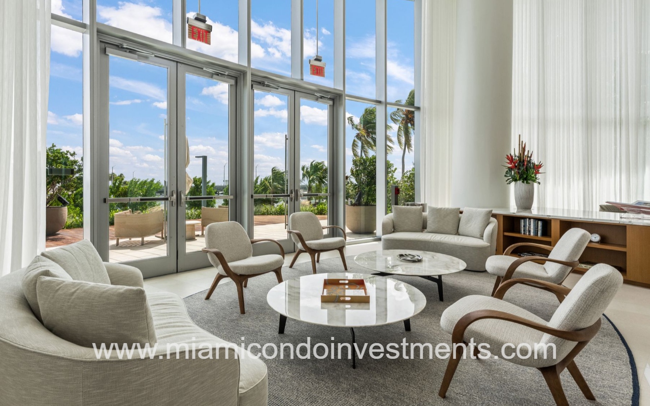 UNA Residences bright hotel lobby or lounge with curved neutral seating around two marble coffee tables, large glass doors to an outdoor patio, and palm trees outside.