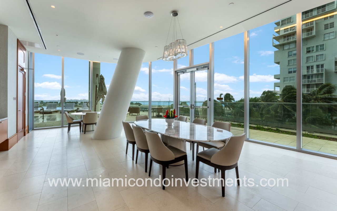 UNA Residences condominium building area with marble table and beige chairs, floor-to-ceiling windows, and Biscayne Bay view.