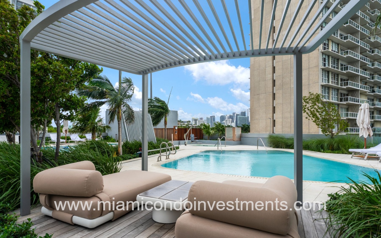 UNA Residences condominium building rooftop pool area with beige cushioned loungers under a metal slatted canopy, palm trees, and a city skyline in the background.